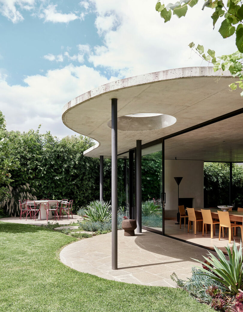 Modern house with a curved concrete roof, large glass walls, and an outdoor dining area surrounded by greenery and a manicured lawn.