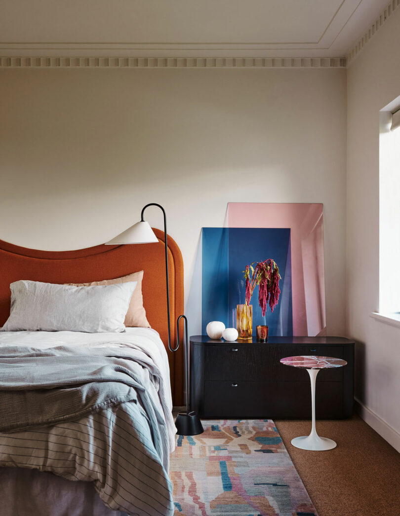 A modern bedroom with an orange upholstered headboard, white bedding, a black nightstand, a tall floor lamp, abstract artwork, and a small round white side table.