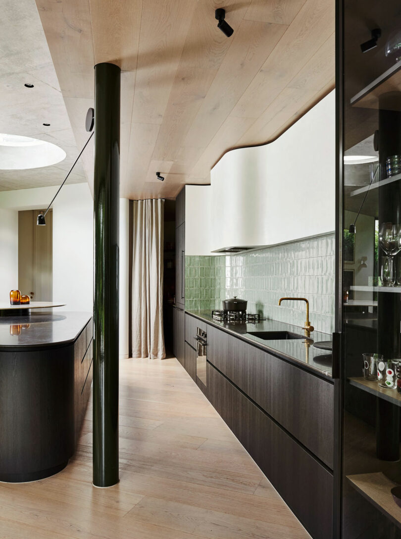 Modern kitchen with dark cabinetry, green tile backsplash, brass faucet, black countertop island, and a glossy green column under a light wood ceiling and floor.