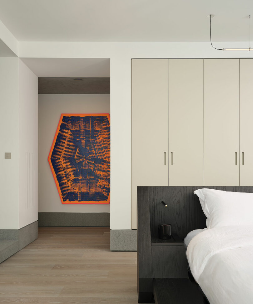 A modern bedroom with light wood flooring, beige built-in wardrobes, a black bed frame, and a geometric orange and blue wall art in the hallway.