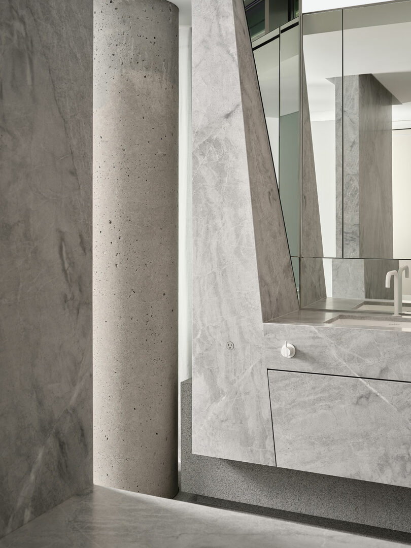 A modern bathroom interior featuring gray stone surfaces, a cylindrical concrete column, angular cabinetry, and large mirrored panels.