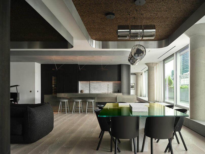 Modern open-concept kitchen and dining area with black chairs, glass table, wood flooring, and large windows offering city views.