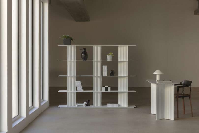 Modern minimalist interior with a white, open shelving unit and desk. Decor includes plants, books, and black-and-white objects. Large windows on the left let in natural light