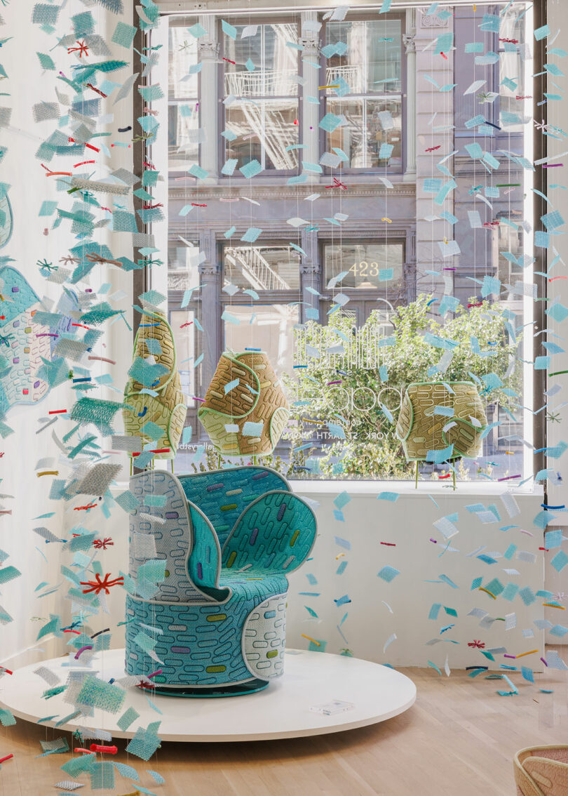 A patterned blue and gray chair is displayed on a round platform in a bright window, surrounded by suspended confetti-like decorations, with buildings visible outside.
