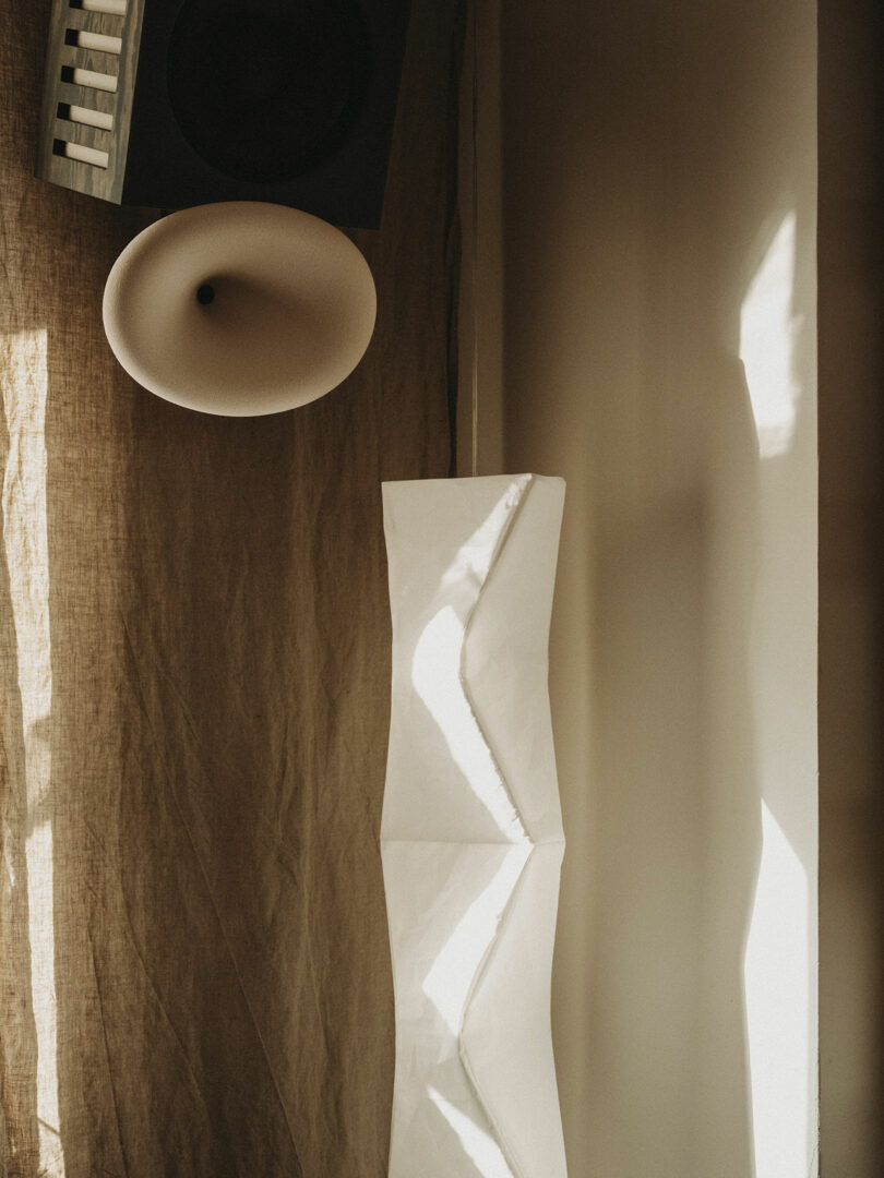 A speaker and a sculptural white lamp are positioned in a corner with textured beige curtains and sunlight casting shadows on the wall.