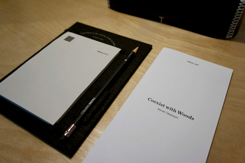 A pencil and notepad are placed on a black felt pad next to a pamphlet titled “Coexist with Woods” by Ryushi Yamashiki, set on a wooden desk