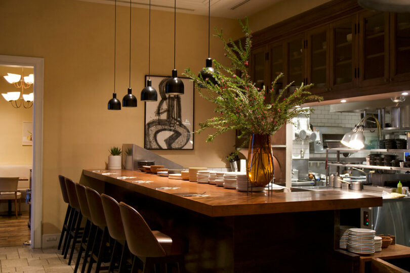 Modern restaurant interior at TRUNK(HOTEL) CAT STREET, featuring a wooden counter, brown chairs, pendant lights, a large vase with greenery, and an open kitchen in the background