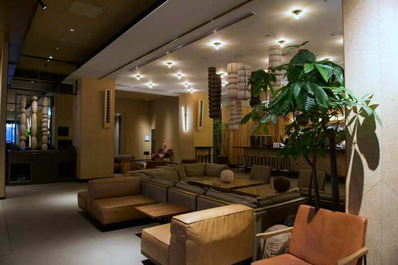 Modern lounge area at TRUNK(HOTEL) CAT STREET with tan leather sofas, armchairs, a large potted plant, hanging light fixtures, and a bar in the background. The space is softly lit and offers a chic, contemporary vibe