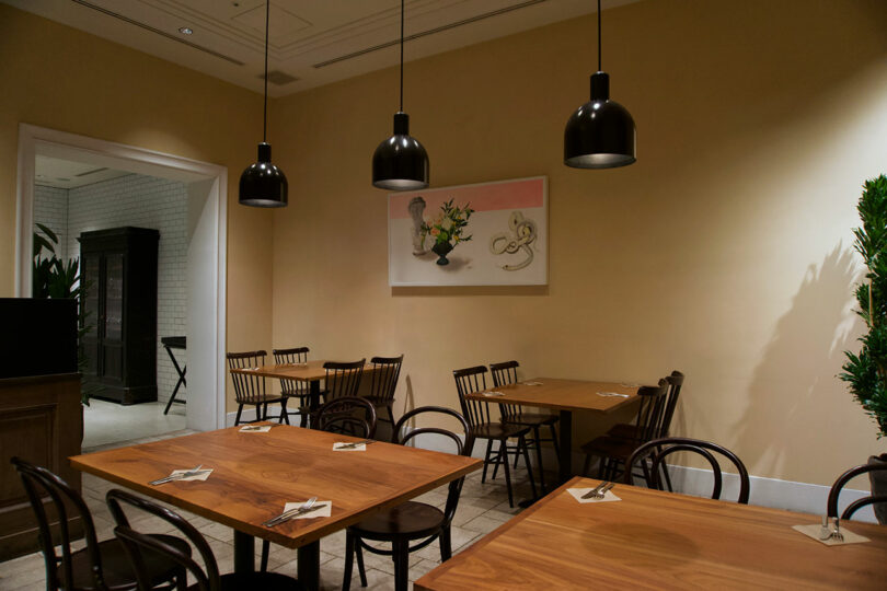 A small, modern dining area at TRUNK(HOTEL) CAT STREET features wooden tables, black chairs, hanging black pendant lights, and a framed artwork on a beige wall