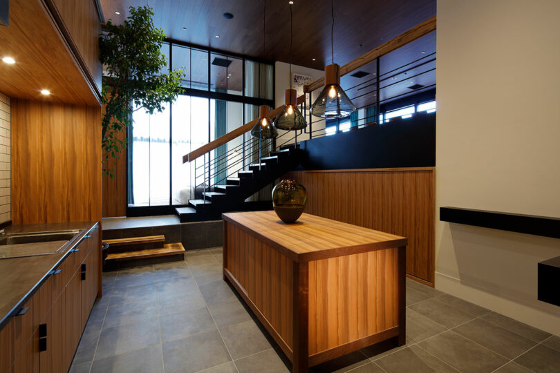 Modern kitchen area in a room at TRUNK(HOTEL) CAT STREET with wood-paneled walls and an island, grey tiled floor, sleek black countertop, large windows, and a staircase with metal railing in the background