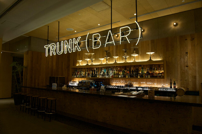 A modern bar with a wooden counter, glassware, and liquor bottles at TRUNK(HOTEL) CAT STREET, featuring a large neon sign reading 