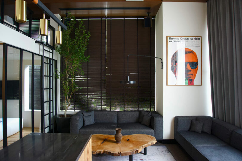 Modern living room with dark gray/navy sofas, a large wooden coffee table, tall plant, window blinds, and a framed poster on the wall featuring a person in sunglasses.