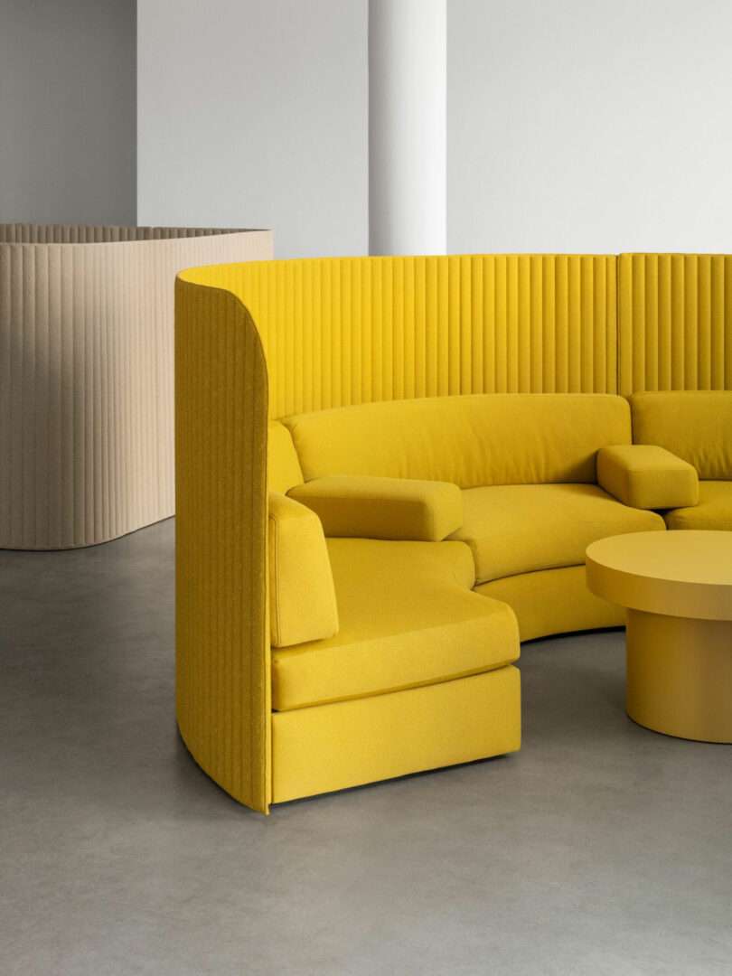 Curved yellow sectional sofa with high ribbed backrest and matching round coffee table in a minimalist, neutral-toned room with concrete floor.