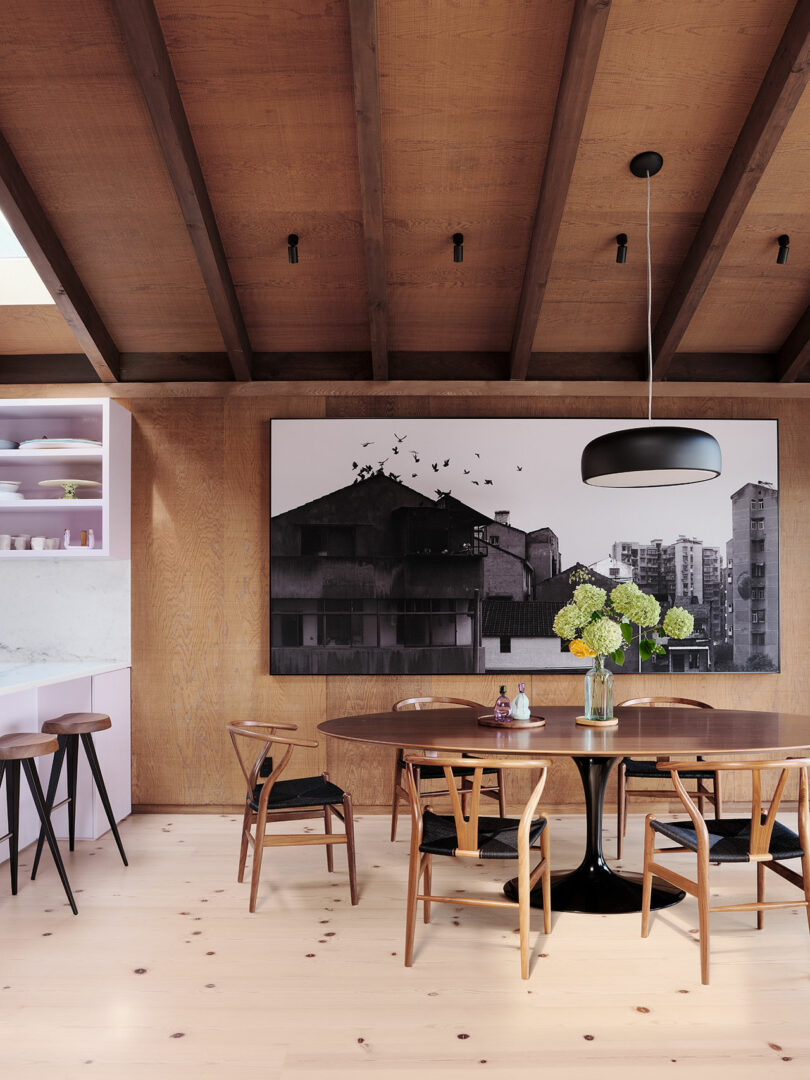 A modern dining area with a round table, wooden chairs, a vase of flowers, a large black-and-white cityscape photograph, and wood-paneled walls and ceiling.