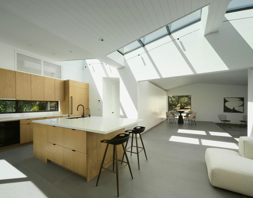Modern kitchen with wood cabinetry, a large island with barstools, and skylights letting in natural light; adjacent dining area and living space visible.