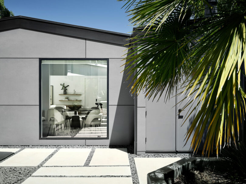 Modern house exterior with large window showing a minimalist interior with white chairs, table, and wall shelves; palm leaves partially obscure the view.