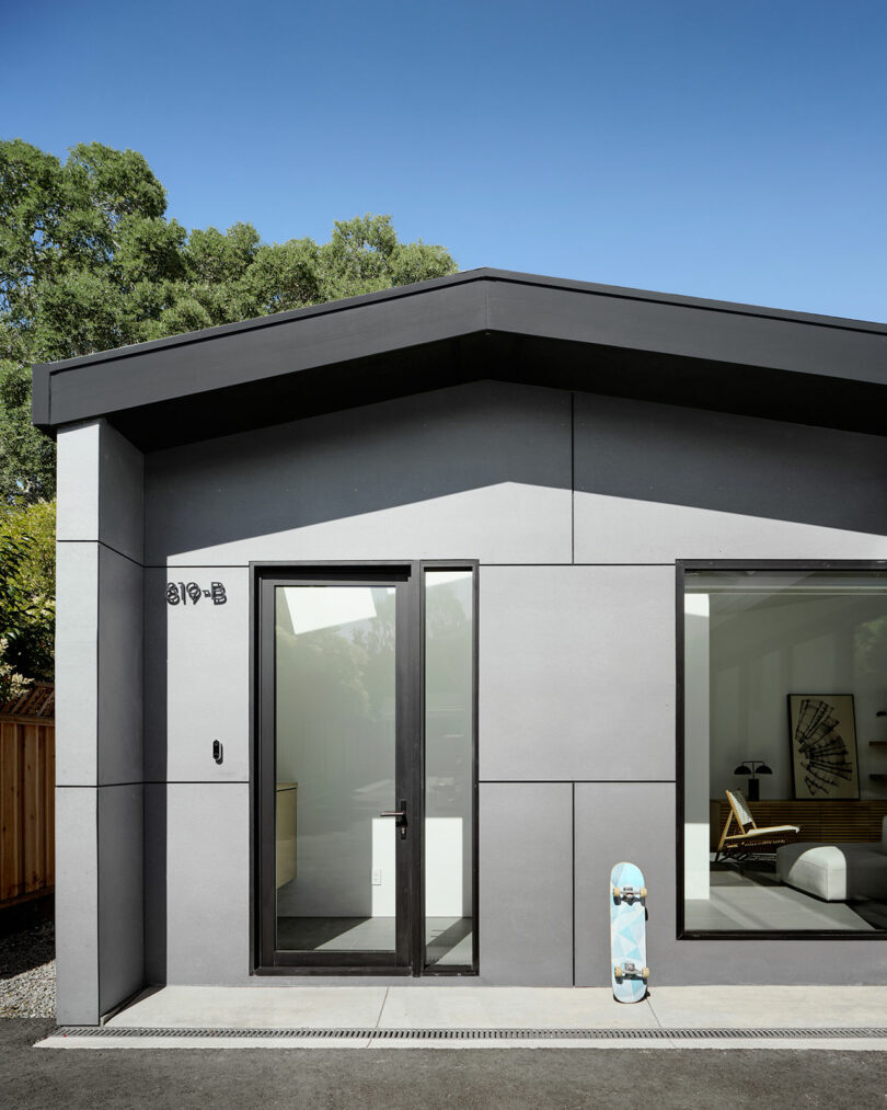 A modern gray house with large windows, a glass door, and a skateboard leaning against the wall on a sunny day. Trees and a wooden fence are visible in the background.