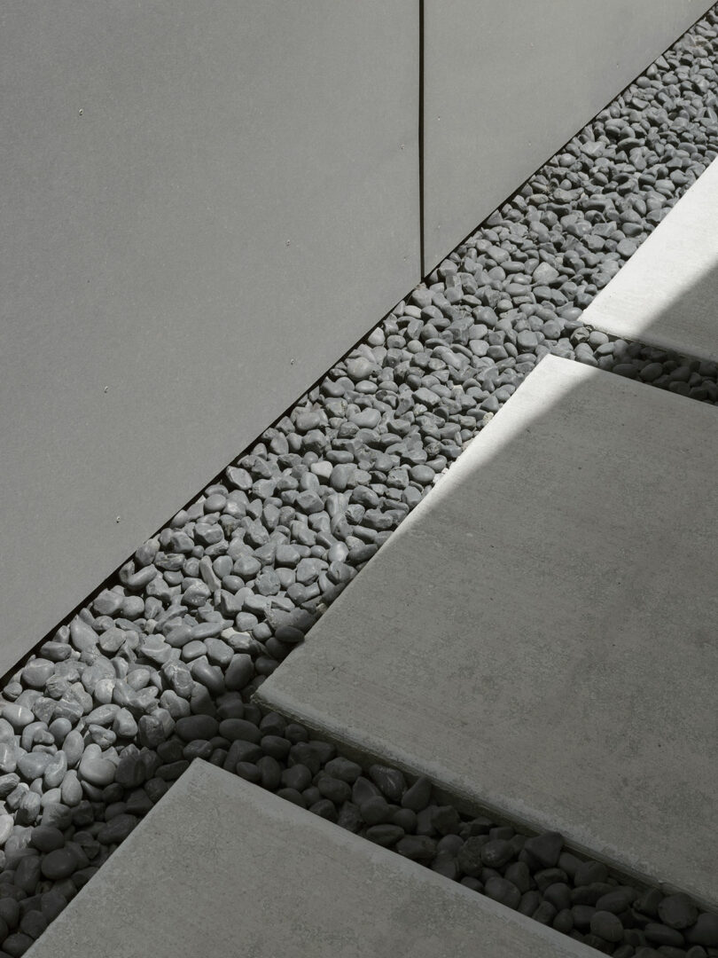 Gray concrete pavers separated by gaps filled with small smooth gray stones, adjacent to a gray wall, with sunlight casting shadows.