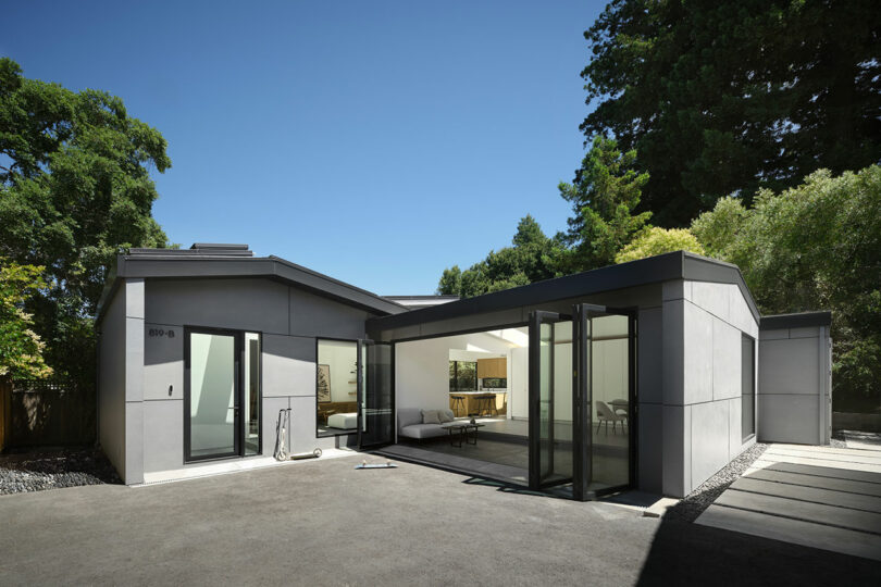 Modern one-story house with large windows and folding glass doors, surrounded by trees, under a clear blue sky.