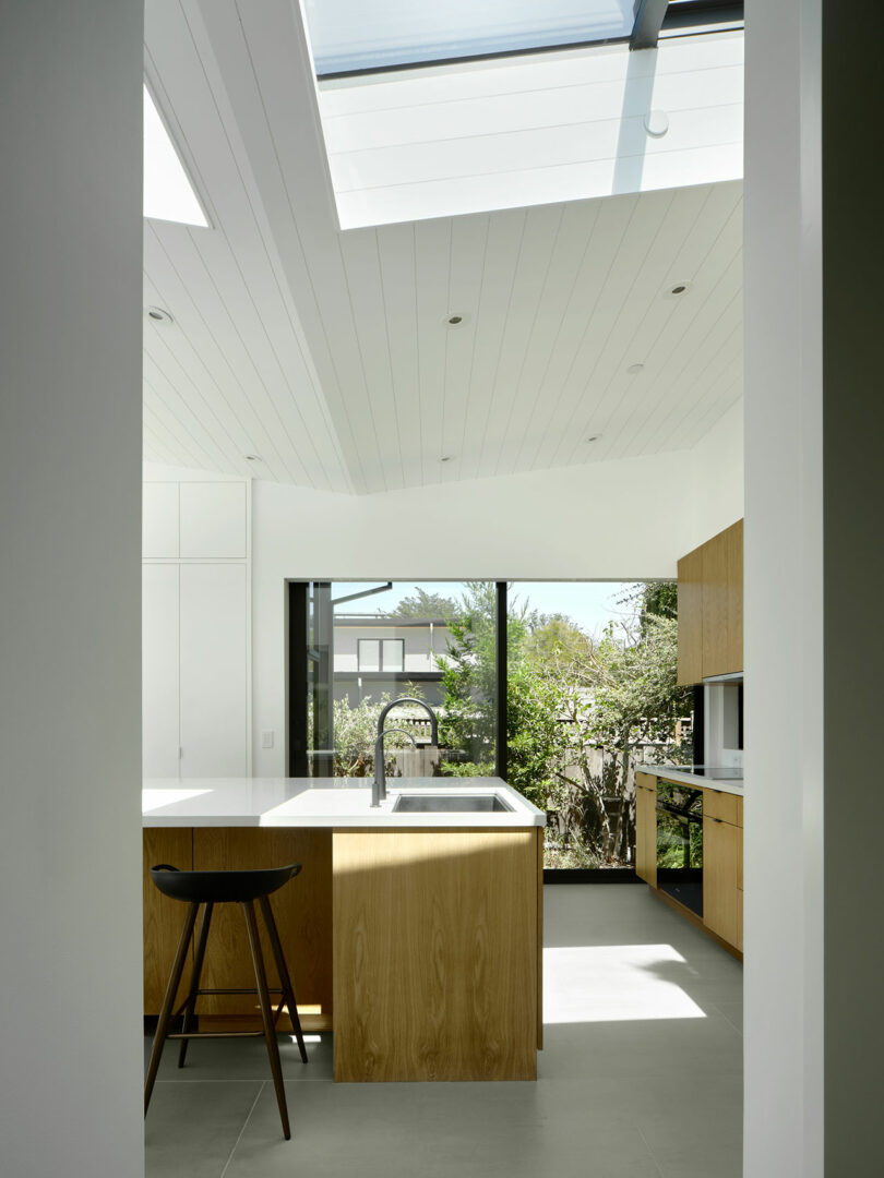 Modern kitchen with wooden cabinets, island with sink, black stool, large windows, and skylights letting in natural light.