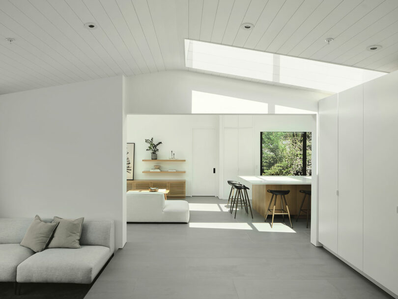 Minimalist living and kitchen area with white walls, light wood furniture, a gray sofa, bar stools at a counter, and large windows letting in natural light.