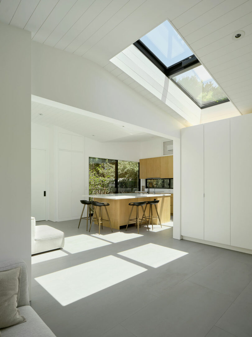 Modern kitchen with wooden island and black bar stools, large windows, skylight, and white walls. Sunlight fills the space, creating a bright and open atmosphere.