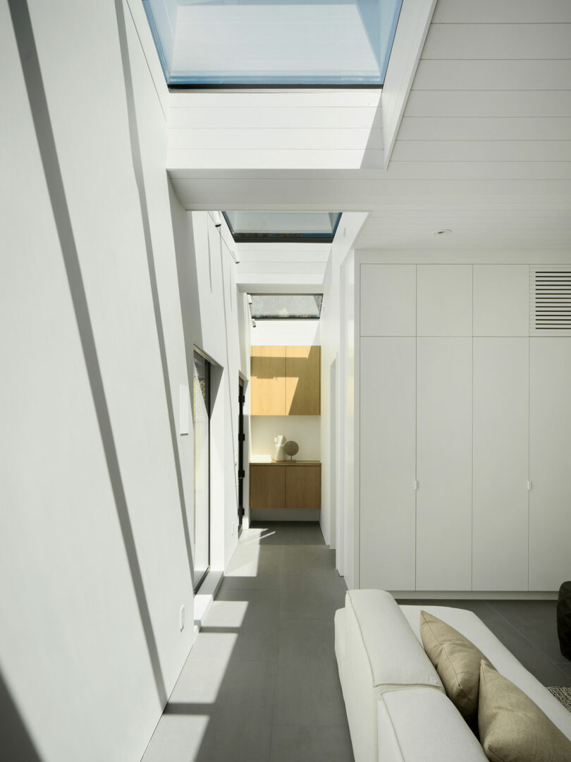 A modern, minimalist interior with white walls, built-in cabinets, a light gray floor, and a skylight allowing sunlight to stream in. A sofa with beige pillows is in the foreground.