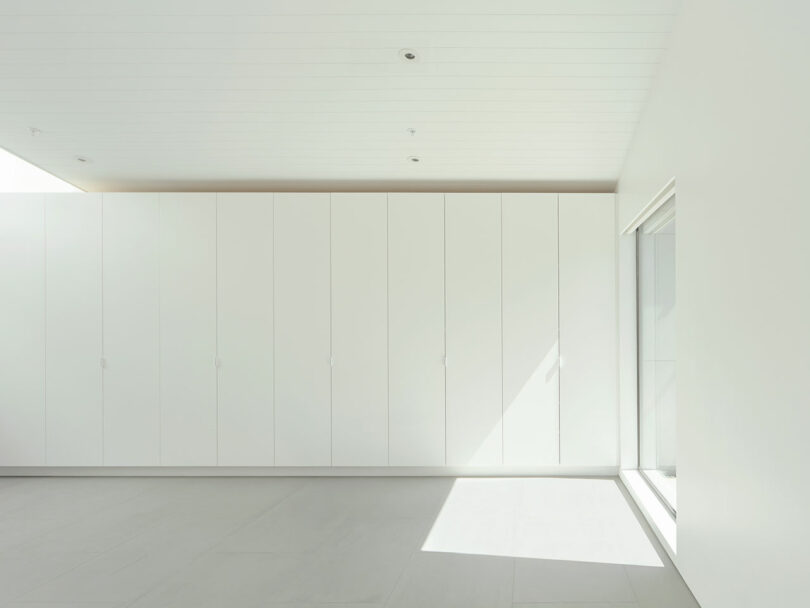Minimalist white room with a wall of floor-to-ceiling cabinets, white ceiling, light gray floor, and sunlight streaming through a large window on the right.