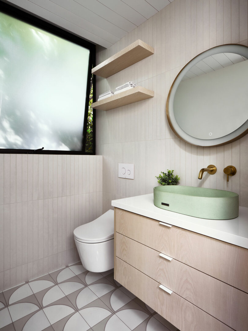 Modern bathroom with a round mirror, green vessel sink, wall-mounted faucet, light wood vanity, open shelves, frosted window, and patterned floor tiles.
