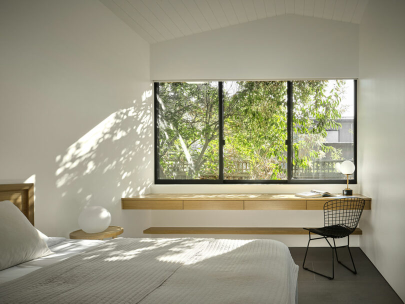 Minimalist bedroom with a bed, floating wooden desk, black chair, and large window overlooking green trees; sunlight casts shadows on the white walls and bedding.