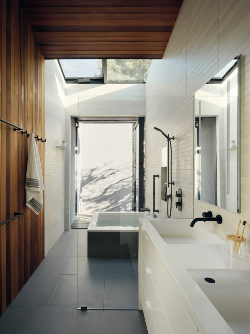 Modern bathroom with white fixtures, wood panel walls, glass shower, soaking tub, large mirror, black fittings, skylight, and natural light coming through windows and ceiling.