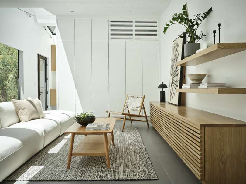 Minimalist living room with a white sofa, woven chair, wooden coffee table, wall shelves, and sideboard; neutral decor with natural light and green plant accents.