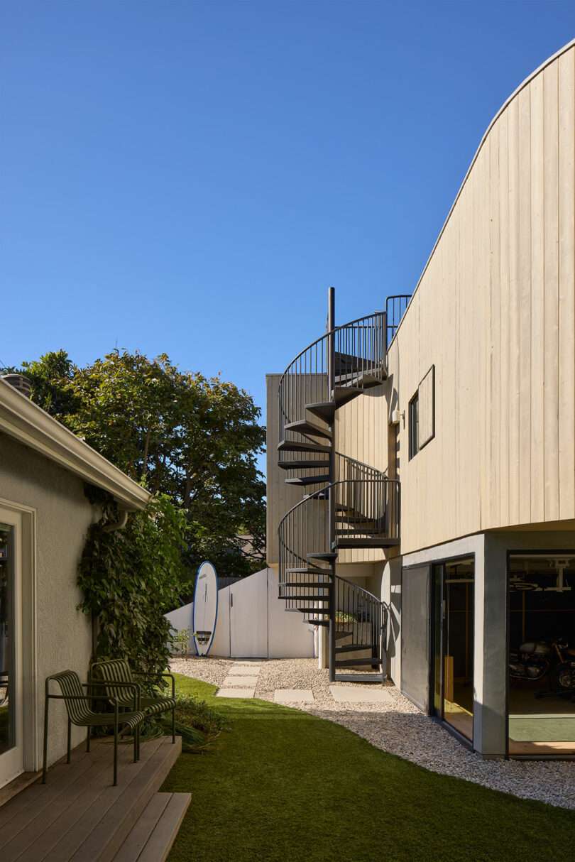 A modern building with light wood siding features an exterior metal spiral staircase, adjacent to a lawn and another structure, under a clear blue sky.