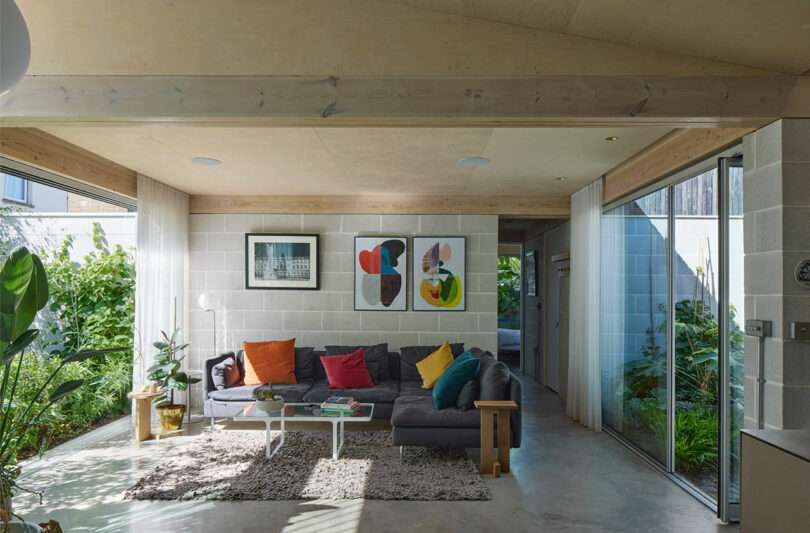 A modern living room with a gray sectional sofa, colorful cushions, abstract art on a white brick wall, plants, a rug, and large windows overlooking greenery.