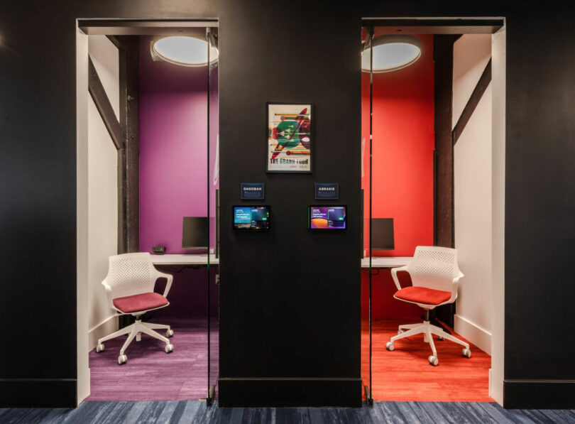 Two small office cubicles with modern chairs and desks, one with purple walls and carpet, the other with red, separated by a black wall, each with a digital room booking panel.