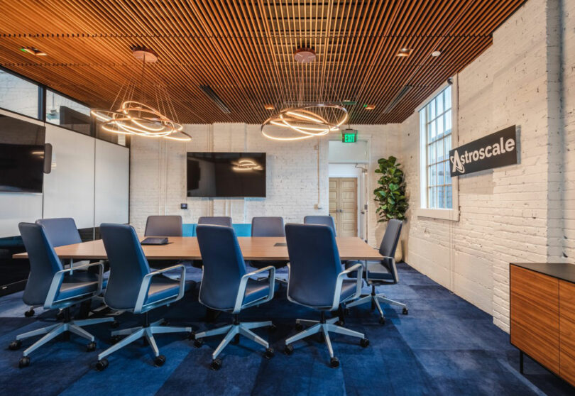 Modern conference room with a long table, blue chairs, wall-mounted screens, geometric light fixtures, and a sign reading 