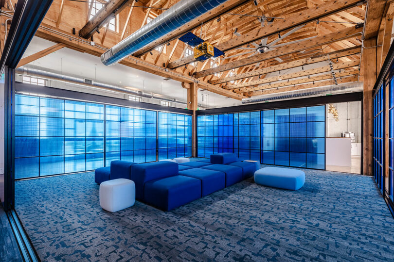 Open office lounge with modular blue seating, Astroscale-inspired blue-tinted glass walls, exposed wooden beams, and industrial ceiling features.