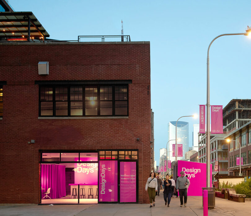 People walk past a brick building and bright pink signs for 