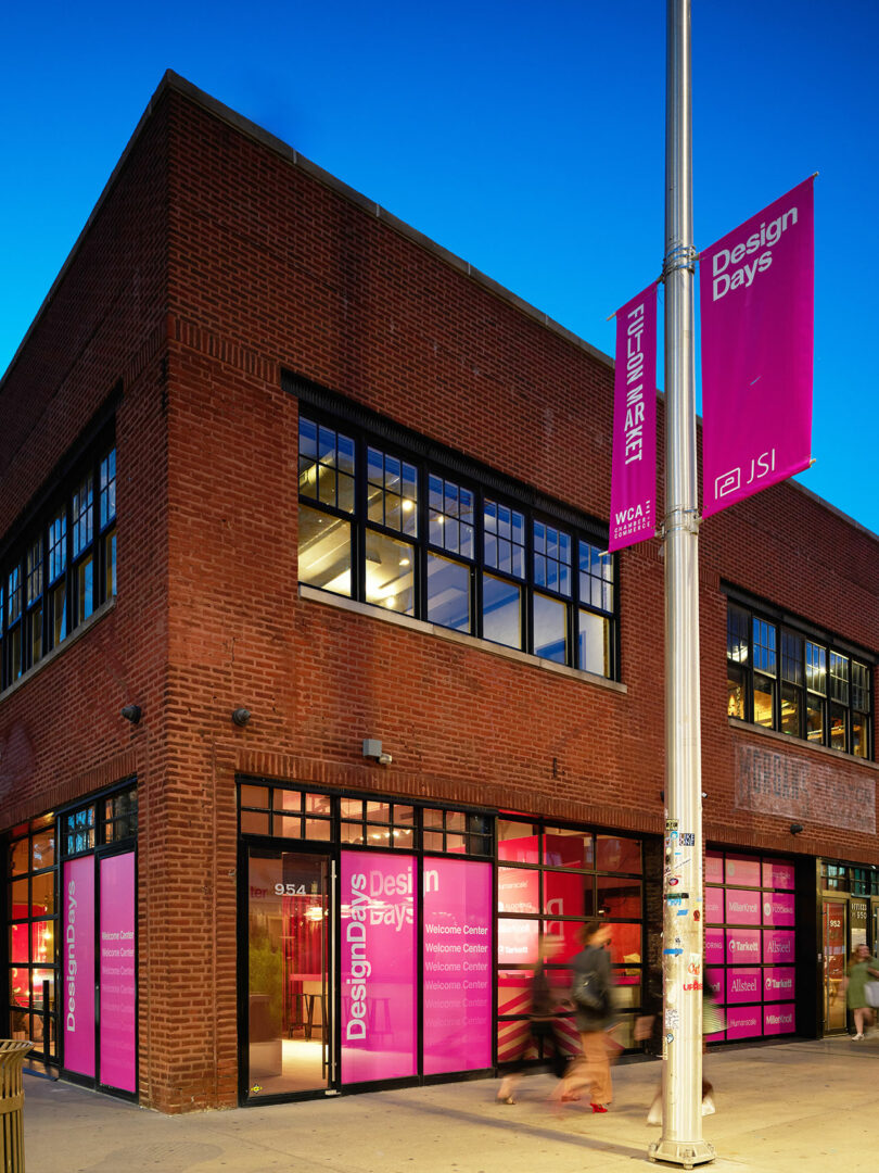 A two-story red brick building with large windows and pink banners reading 