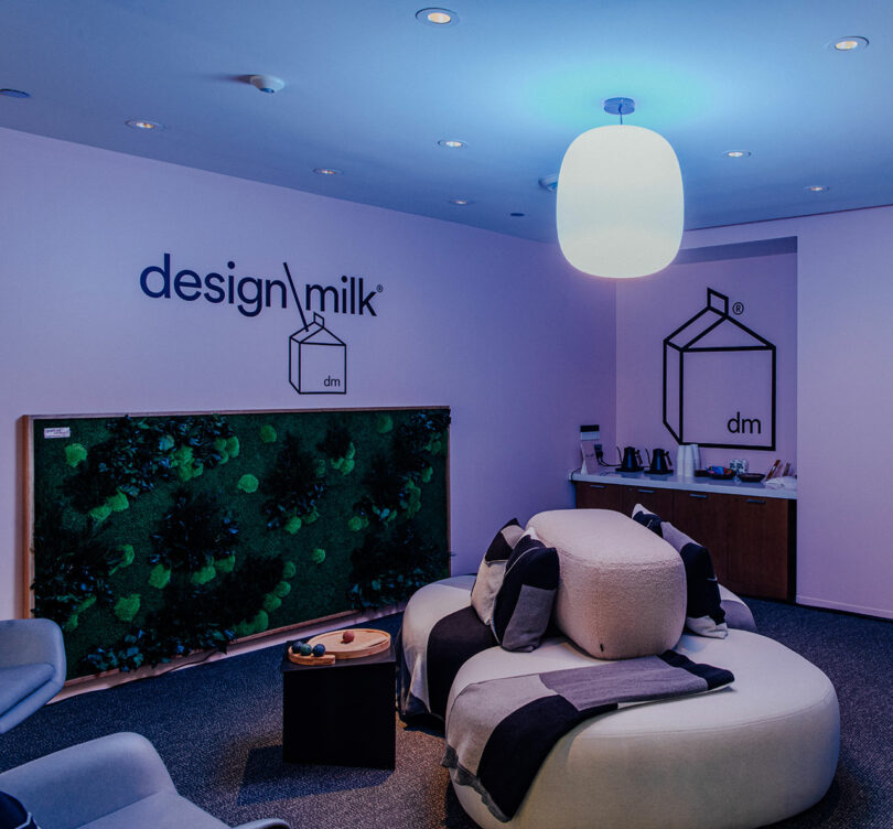 Modern Designer Wellness Lounge with round sofa, pillows, a coffee table, wall art, a plant panel, and Design Milk logos on the walls under soft blue lighting.