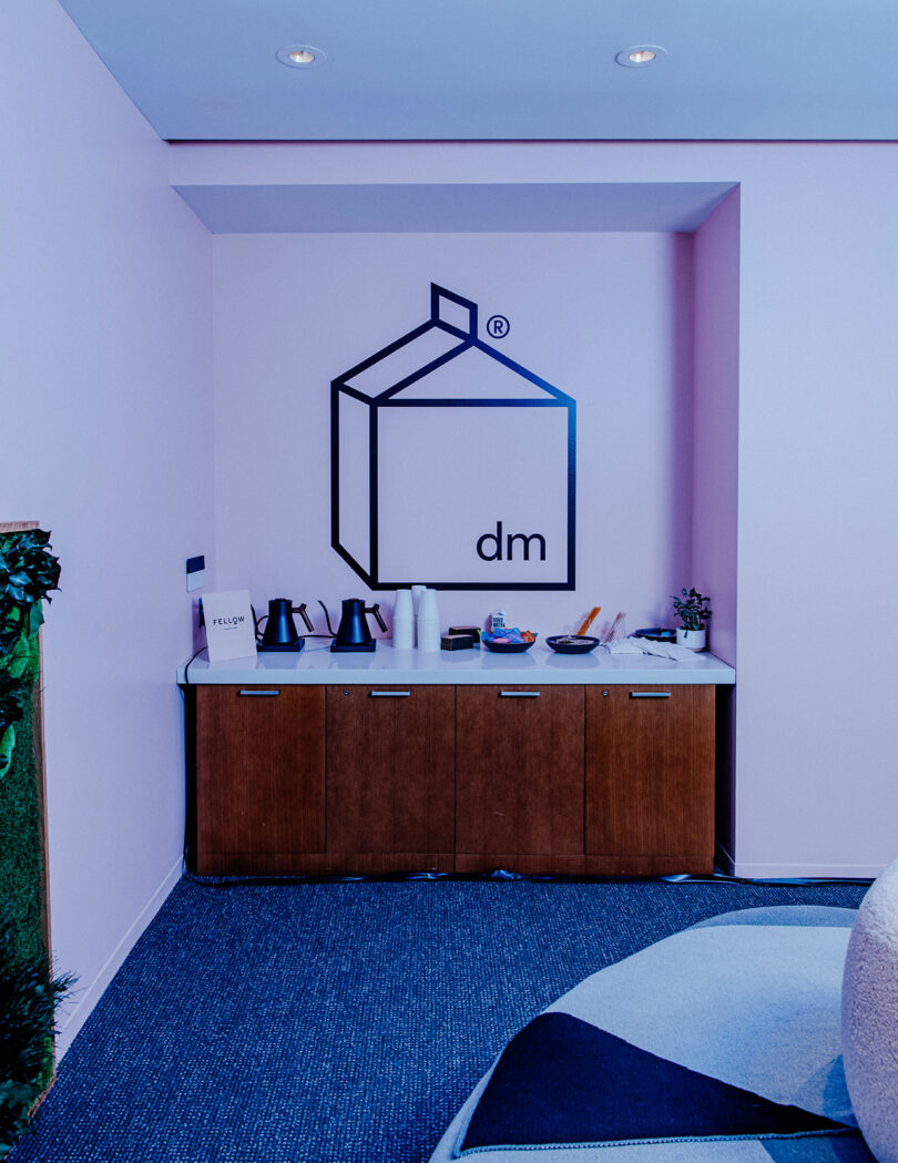A small refreshment station with kettles, cups, and snacks sits against a pink wall with a geometric logo and the letters 