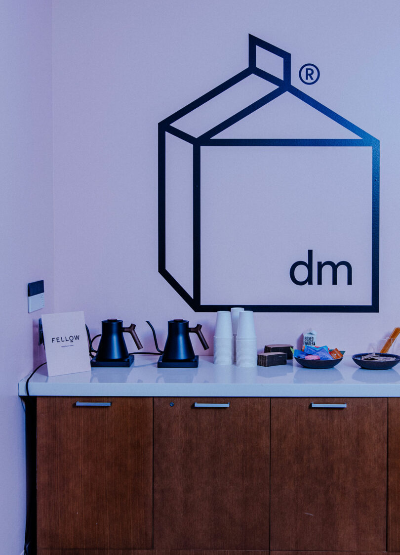 A countertop with two electric kettles, stacked white cups, snacks, and a “Fellow” box creates a stylish refreshment area; above, the geometric “dm” logo hints at a Designer Wellness Lounge vibe.