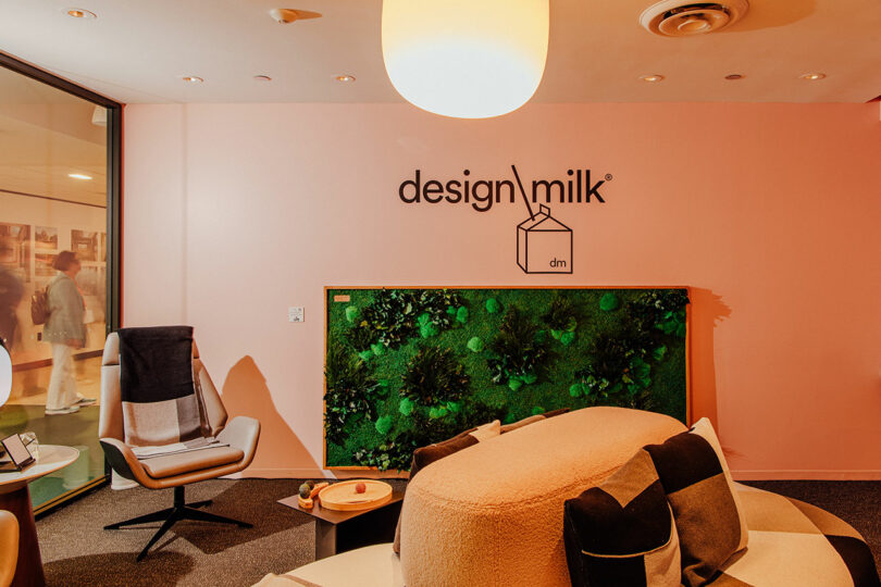 A modern Designer Wellness Lounge with pink walls, a Design Milk logo, a lush green plant wall feature, and contemporary seating including a chair and sofa beneath a ceiling light.