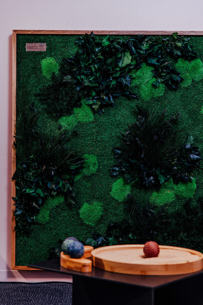 A framed green moss wall art hangs on a white wall in this Designer Wellness Lounge, behind a wooden tray table holding two decorative spheres.