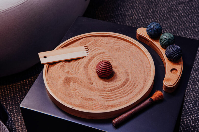A tabletop sand garden with a wooden rake, patterned balls, and a brush arranged on a black surface—perfect for adding calm to your Designer Wellness Lounge.