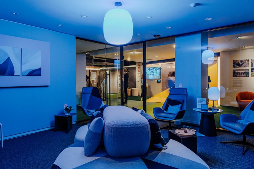Modern Designer Wellness Lounge featuring blue ambient lighting, contemporary chairs, a large cushioned ottoman, and glass walls that reveal a corridor and meeting rooms beyond.