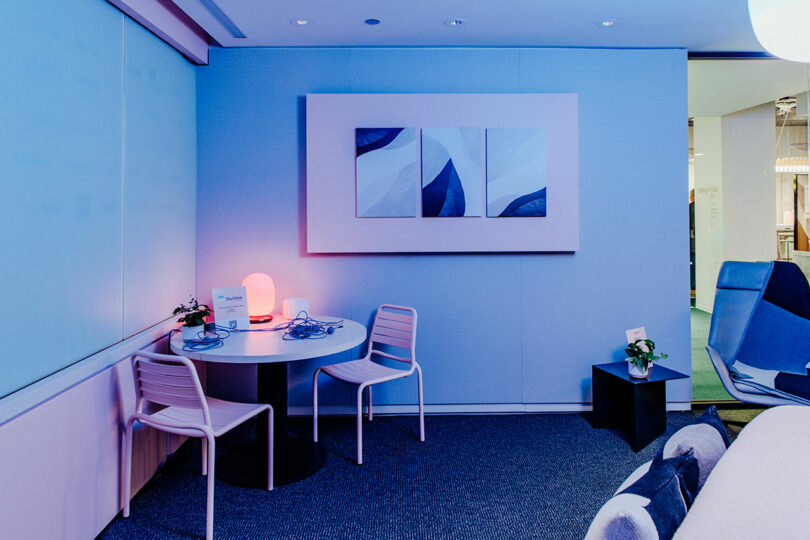 A small modern office corner, designed as a Designer Wellness Lounge, features a round table, two chairs, wall art, and a plant-topped side table, all softly illuminated by blue and pink light.