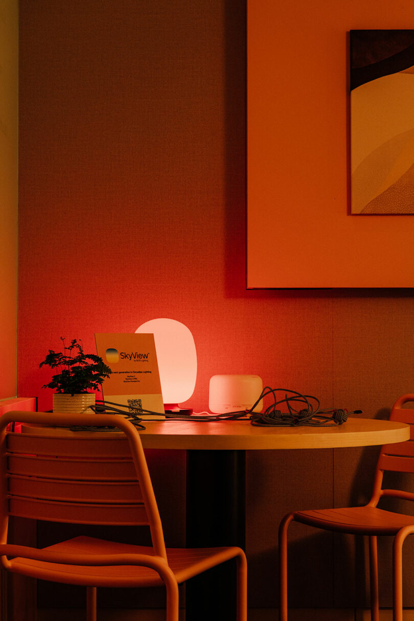 A round table with two chairs, a potted plant, some papers, a lit lamp, and electronic cables sits under warm orange lighting beside abstract wall art in this Designer Wellness Lounge.