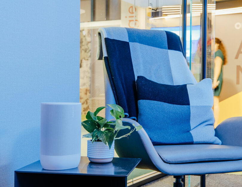 A modern Designer Wellness Lounge setup featuring a gray chair draped in a blue checkered blanket, a small potted plant, and a white Sonos speaker on a sleek black table.