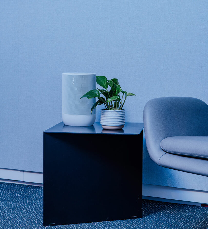 A white smart speaker and a potted plant sit on a black square table next to a gray chair against a blue wall, creating a cozy Designer Wellness Lounge vibe.
